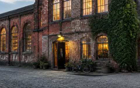 Glowing arched windows, an illuminated entrance and a parked bicycle outside a converted red brick industrial building